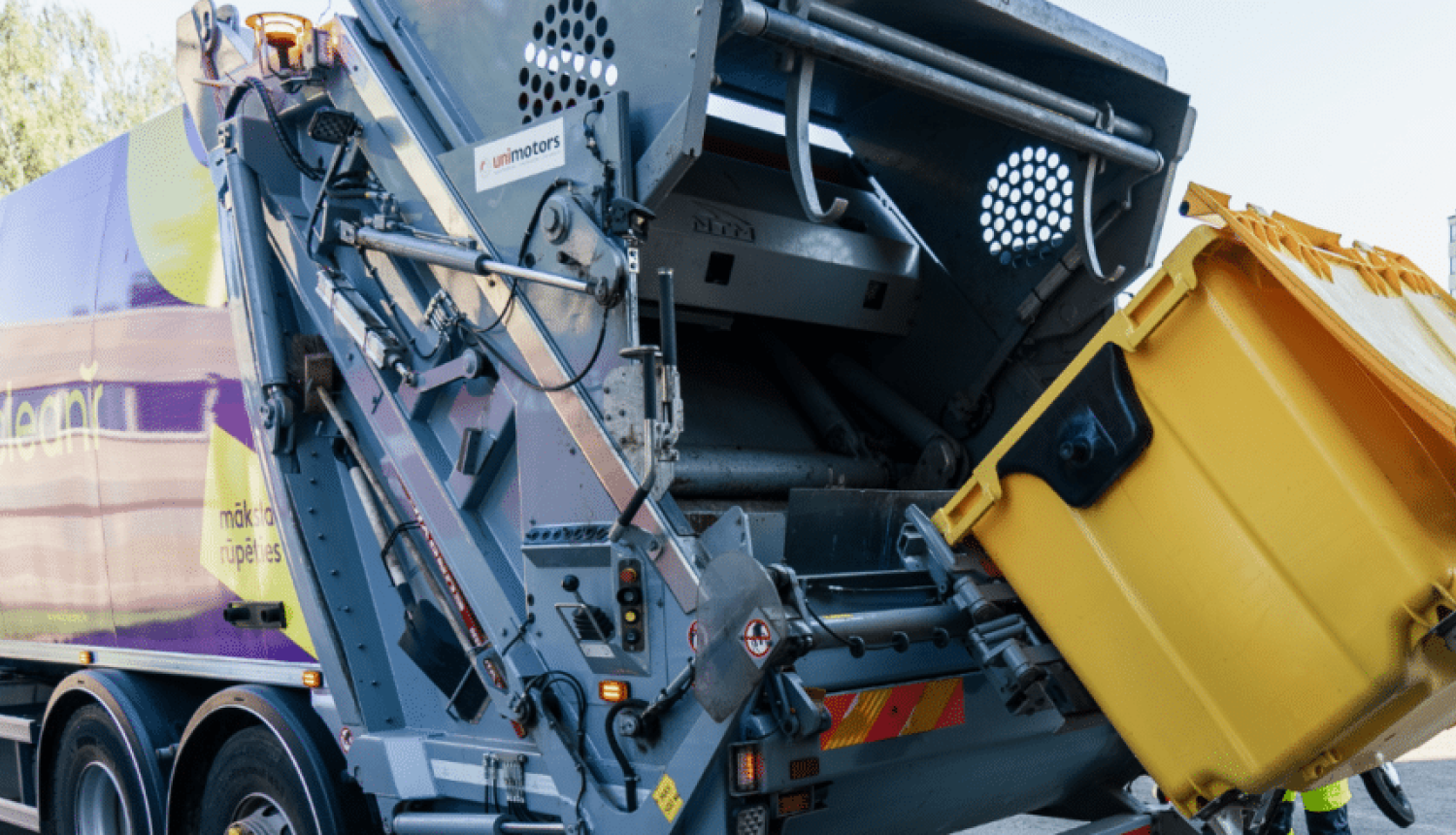 Atkritumu savākšanas mašīna paceļ un iztukšo lielu dzeltenu konteineru