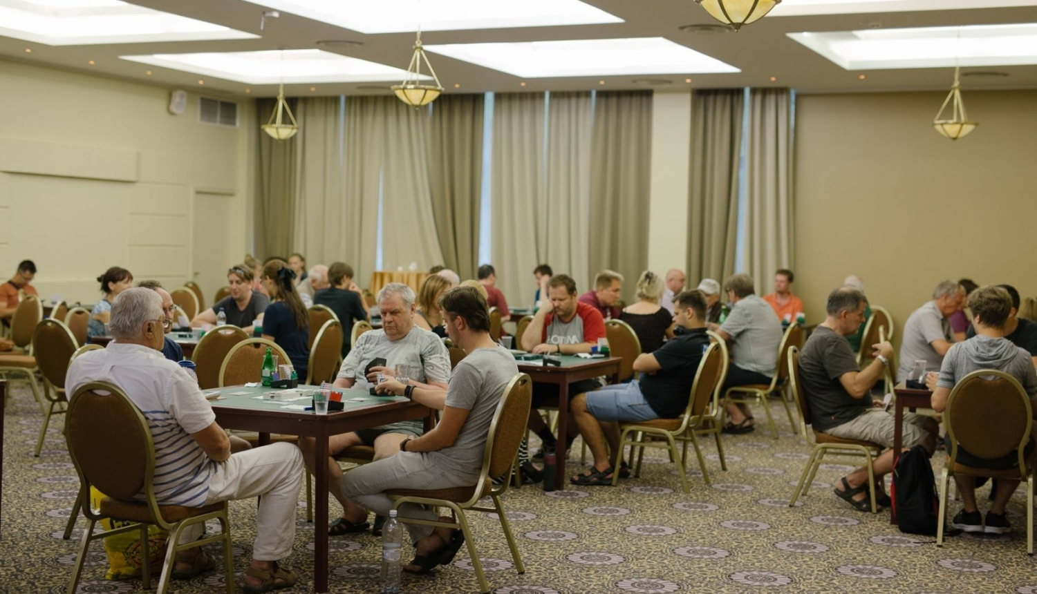 Cilvēki telpā spēlē bridžu pie vairākiem galdiņiem