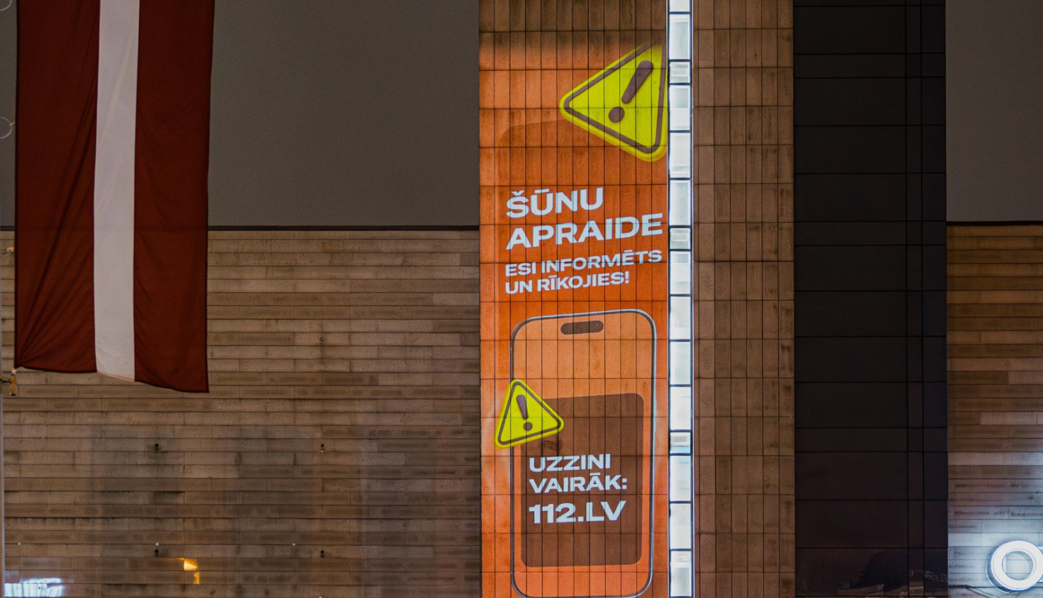 Ēkas fasāde ar izgaismotu vertikālu reklāmu oranžā krāsā, kurā redzams brīdinājuma simbols un teksts latviešu valodā: ‘Šūnu apraide. Esi informēts un rīkojies! Uzzini vairāk: 112.lv