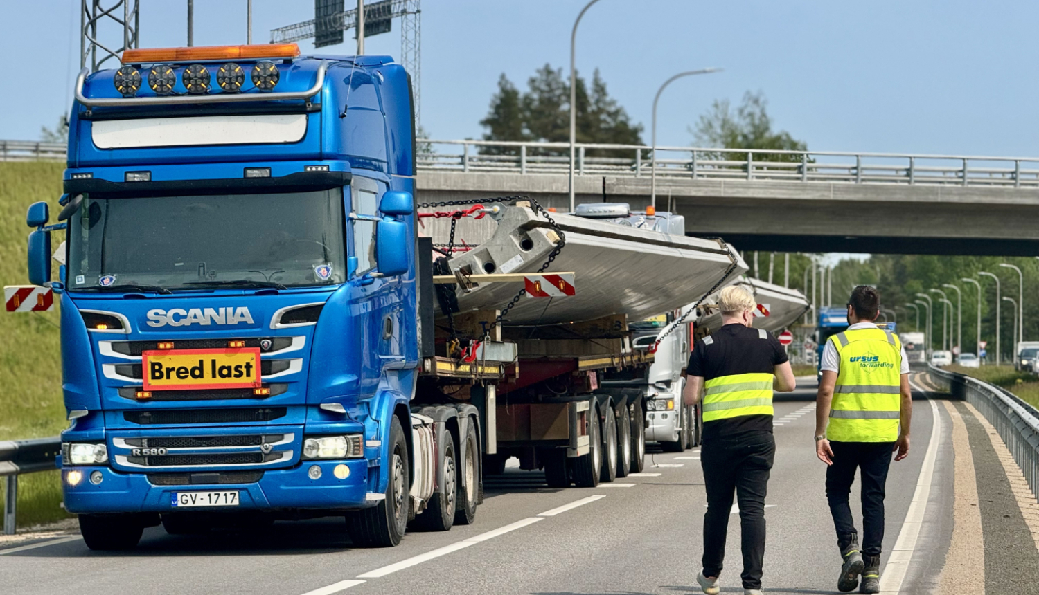 Attēlā redzama zila Scania kravas automašīna ar piekabi, kas pārvadā lielu, platu kravu – iespējams, metāla vai betona konstrukciju. Kravas priekšā ir dzeltena zīme ar sarkanu apmali un uzrakstu "Bred last", kas zviedru valodā nozīmē "Plata krava". Divi vīrieši ar atstarojošām vestēm, viens no kuriem pārstāv uzņēmumu "Ursus Transport", iet gar kravas automašīnu pa ceļa nomali. Fonā redzama automaģistrāle ar vairākām joslām, tilts un citas automašīnas. Attēls ataino kravas pārvadājumu procesa pārraudzību vai
