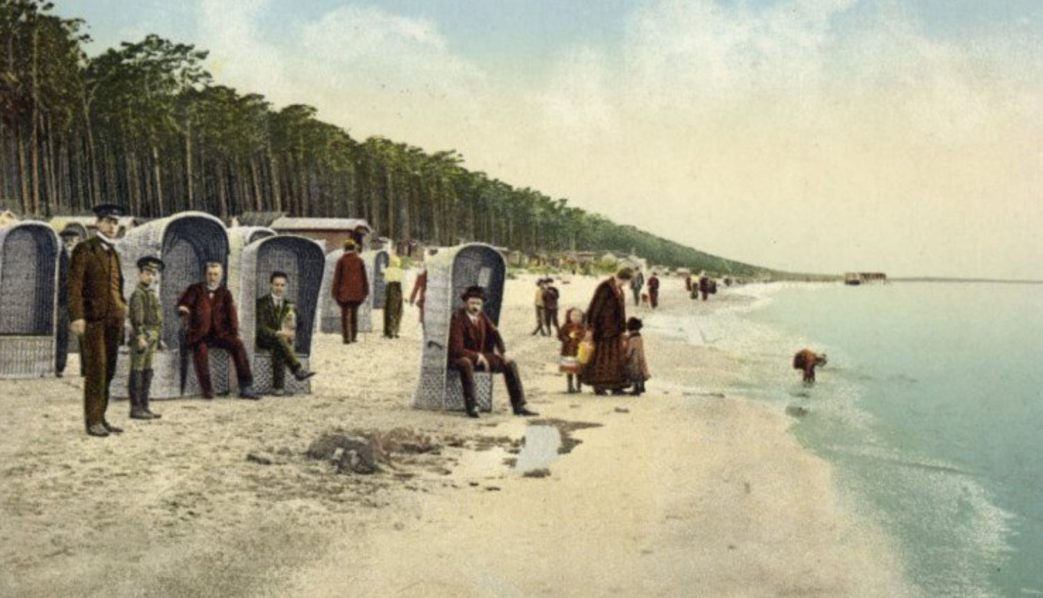 Vēsturiska pastkarte ar cilvēkiem Jūrmalas pludmalē — pie jūras sēž un pastaigājas atpūtnieki, fonā redzama priežu mežs un jūras krasts